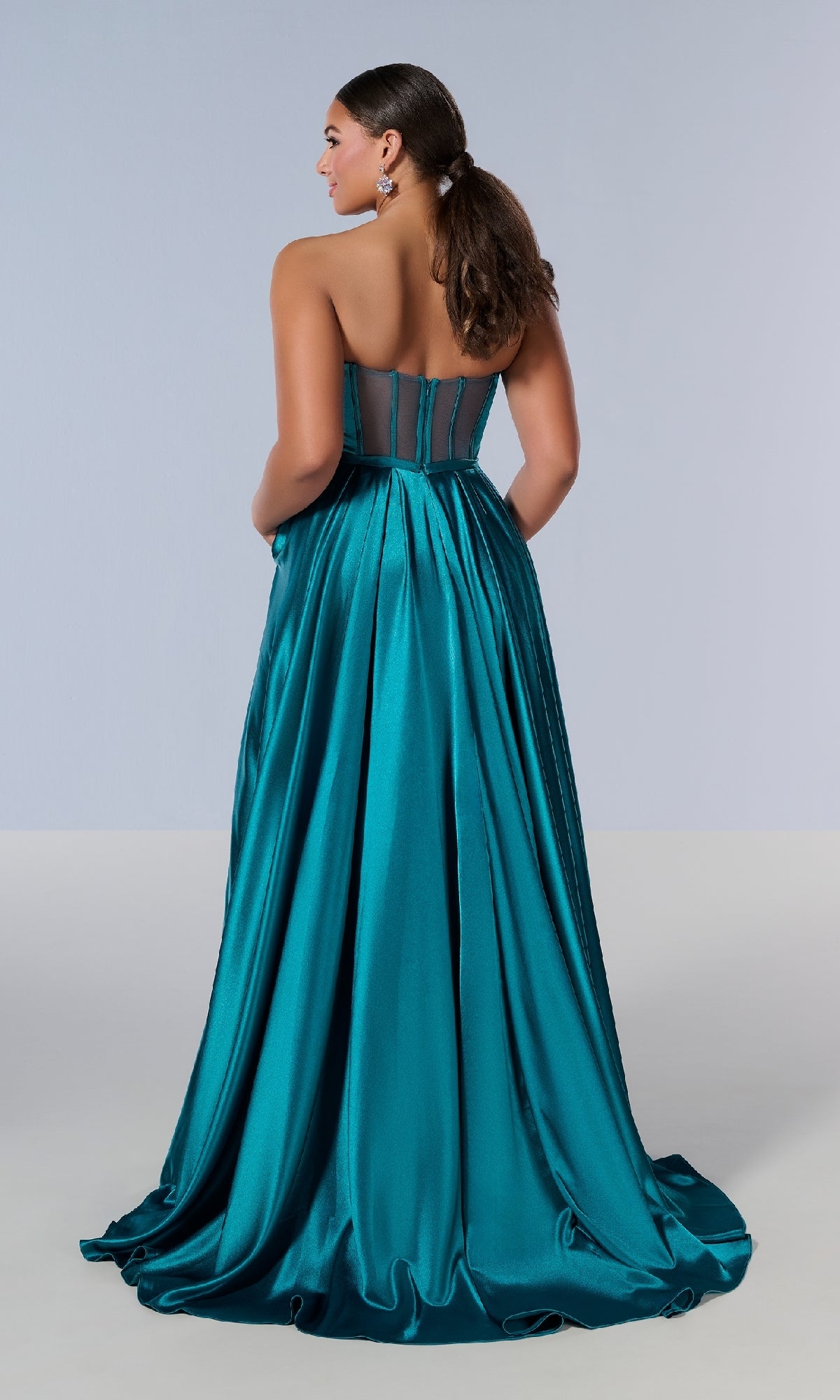 Woman wearing the Long Plus-Size Prom Dress Tiffany 16219 in teal satin, featuring a strapless fitted bodice and sheer back panel, stands with her back to the camera against a plain background.